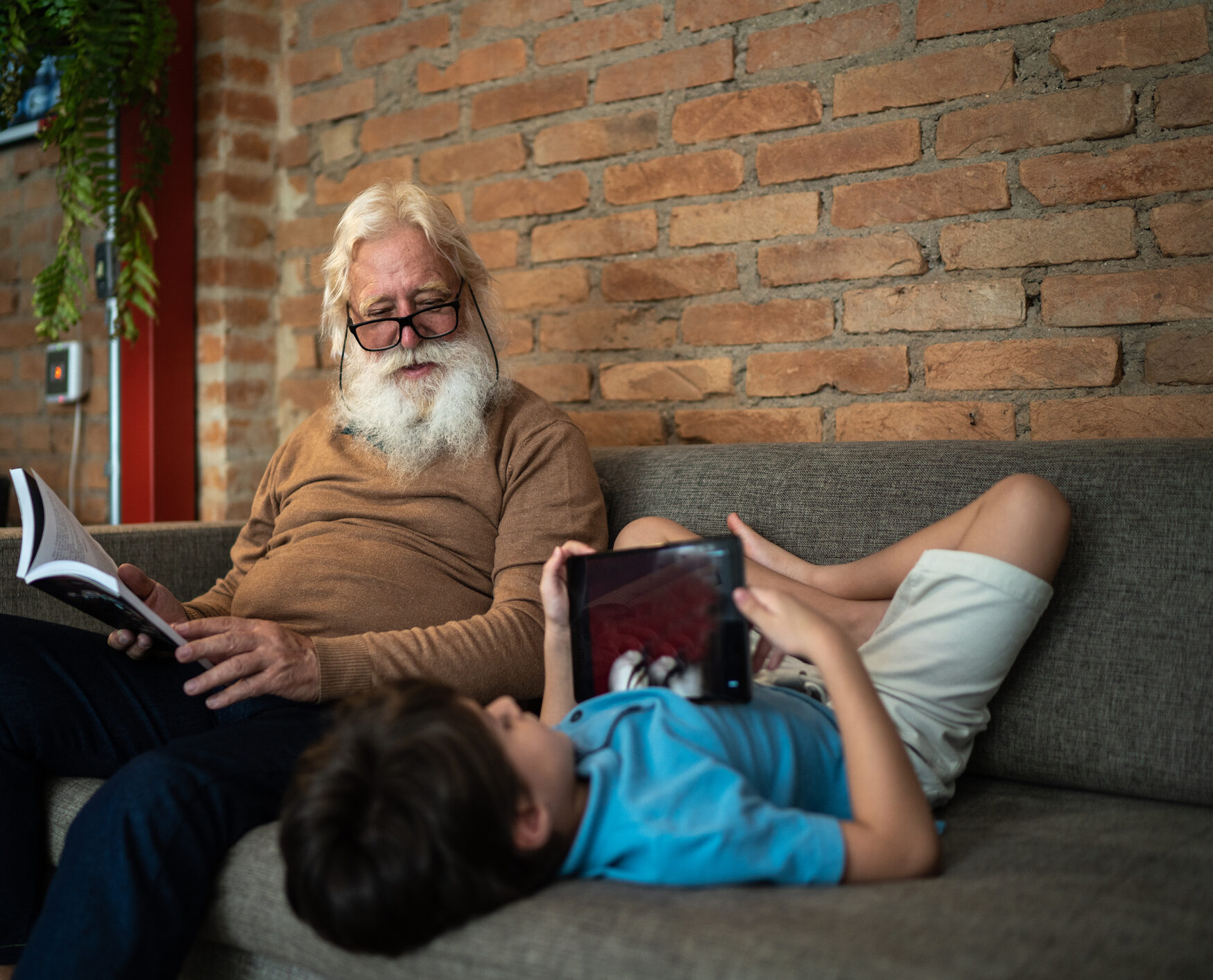 un grand père assis sur son canapé, un livre à la main, regarde son petit fils installé à l'envers les pieds au mur jouant sur sa tablette