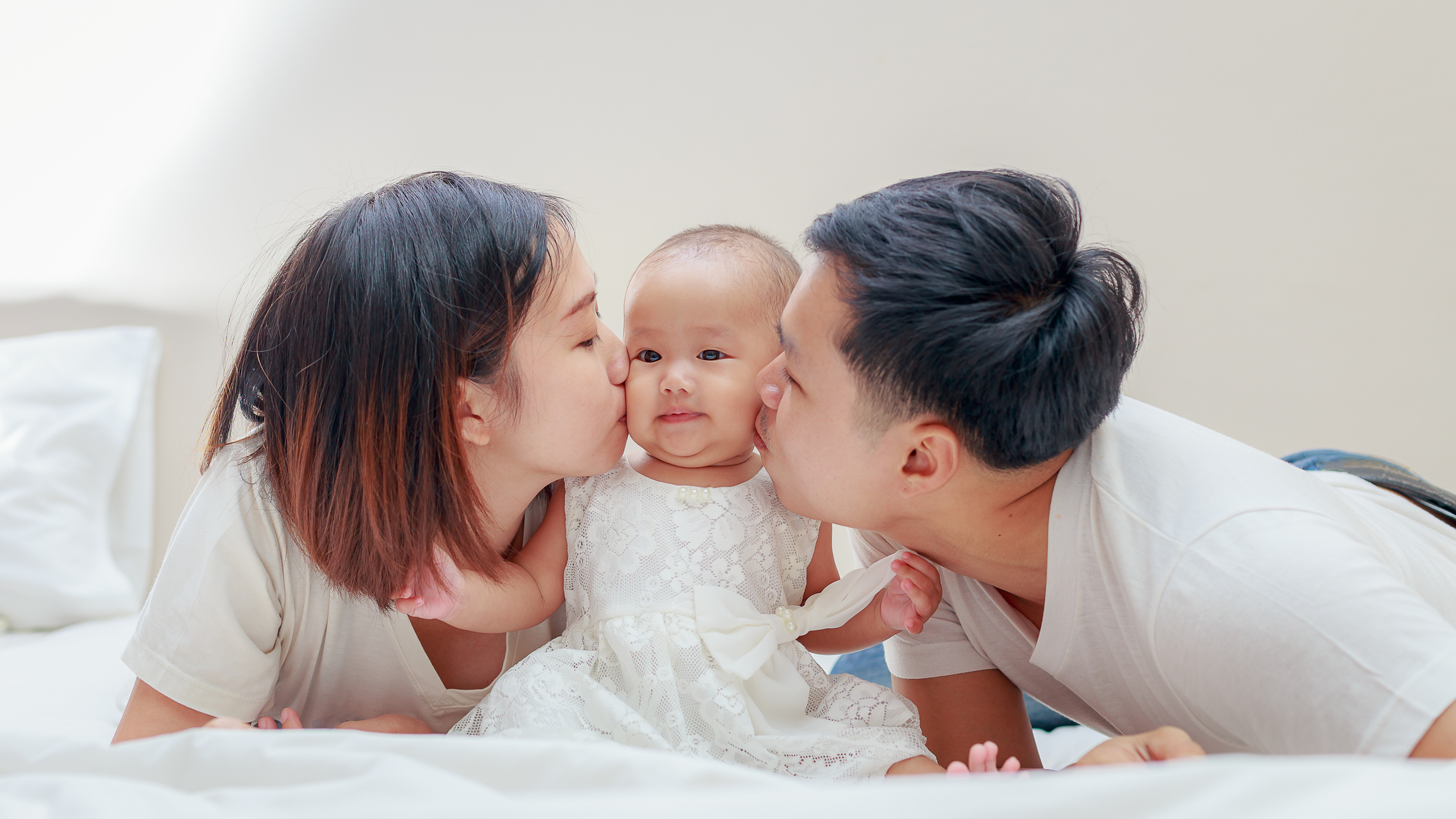 Un couple de jeune parent embrasse leur petite fille habillée d'une petite robe de fête installée sur un lit